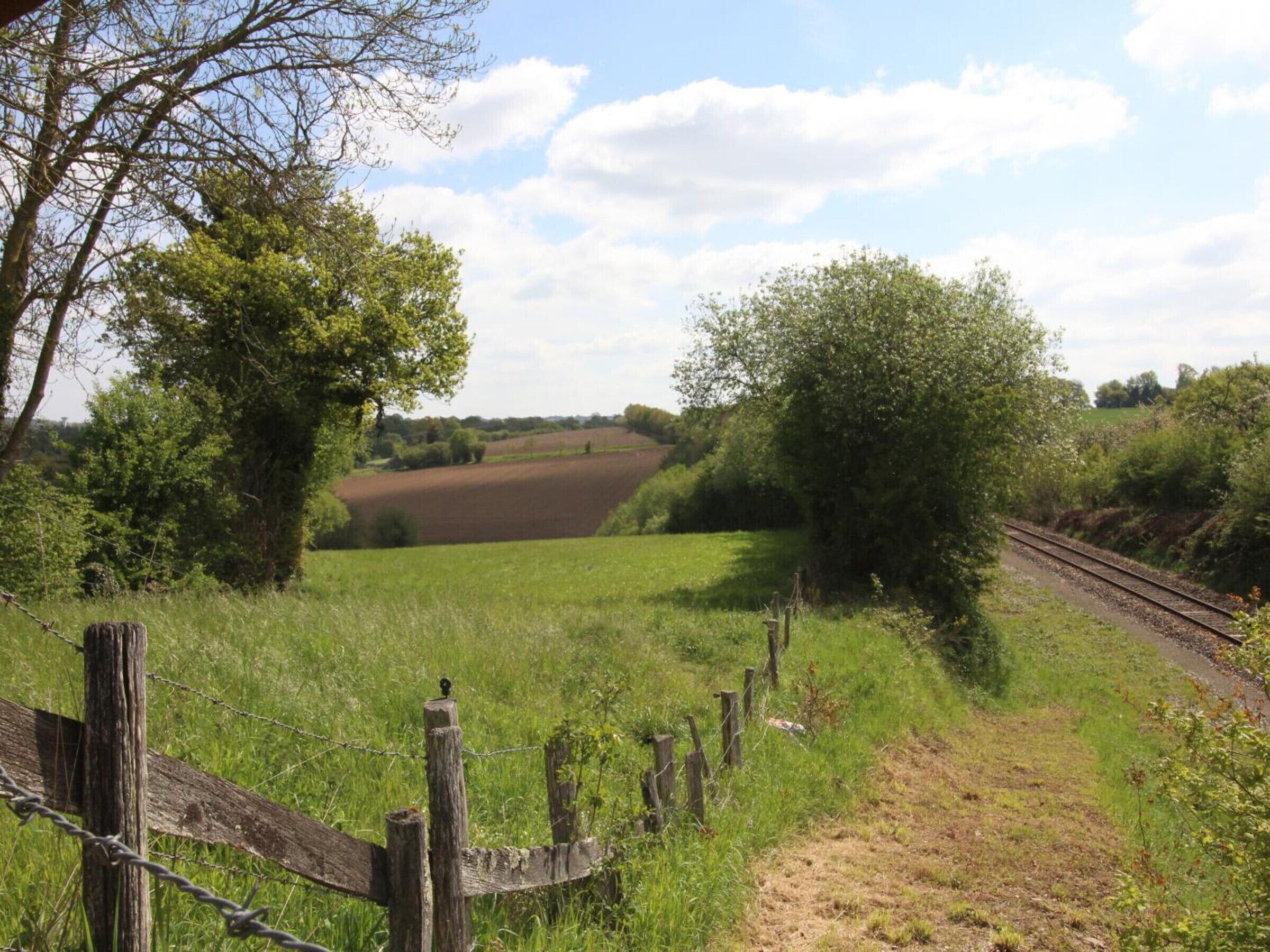 A rural landscape with a dirt path leading to a railway track, surrounded by greenery and fields.