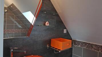 Un bagno dal design moderno, con un lavandino rotondo, uno specchio e uno scaffale in vetro con una piccola scatola arancione.