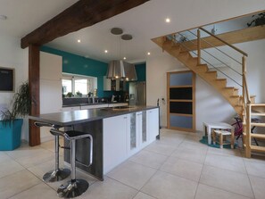 A modern kitchen with a central island, a dining area with a wooden table, and a staircase leading to an upper level. - Ancient house with sauna and games in the heart of Perche (Moussonvilliers)