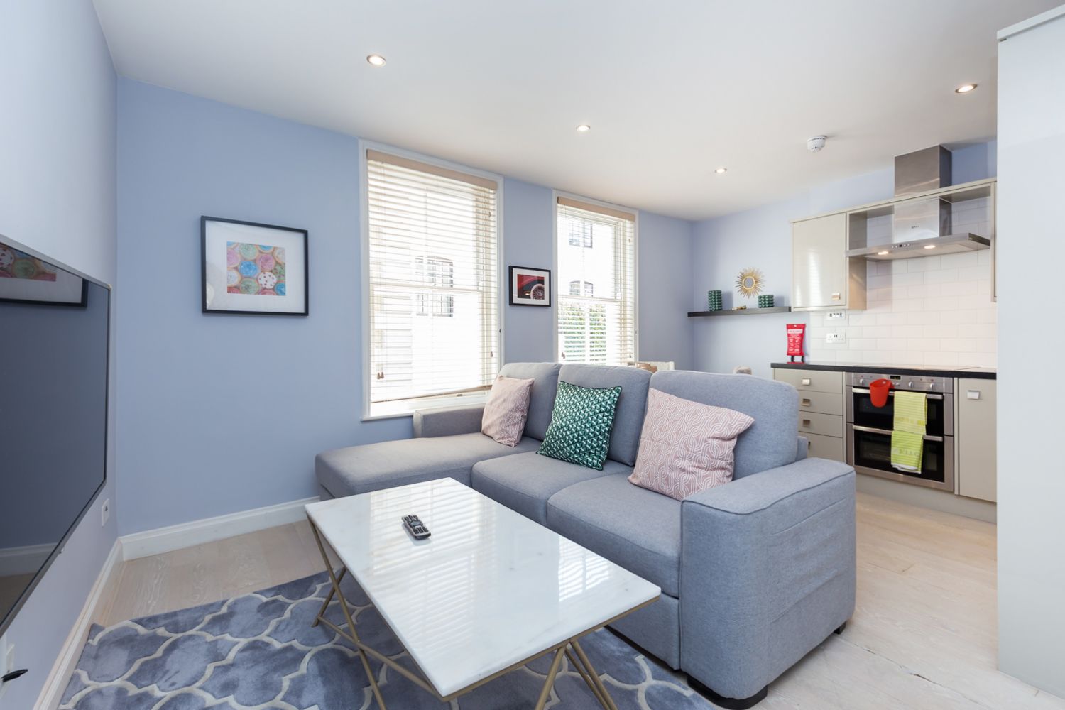 A modern living room with a grey sofa, a white coffee table, and a patterned rug. There are two windows with blinds, a kitchen area with stainless steel appliances, and framed artwork on the walls.