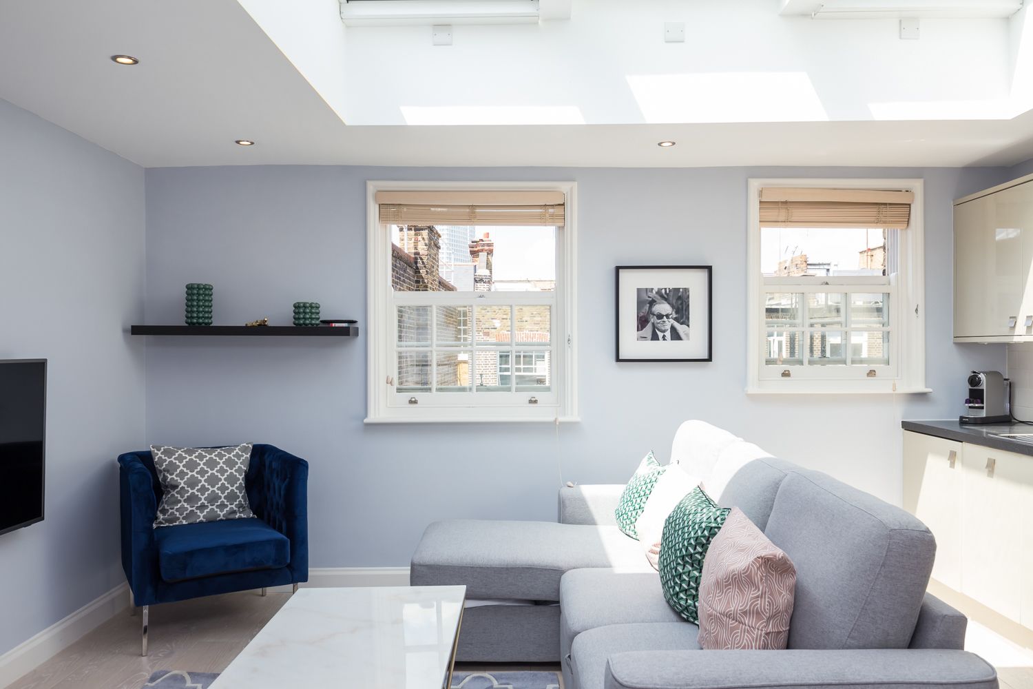 A modern living room with a blue armchair, grey sofa, and a white coffee table. There are two windows with blinds, a framed picture on the wall, and a flat-screen TV mounted on the wall.