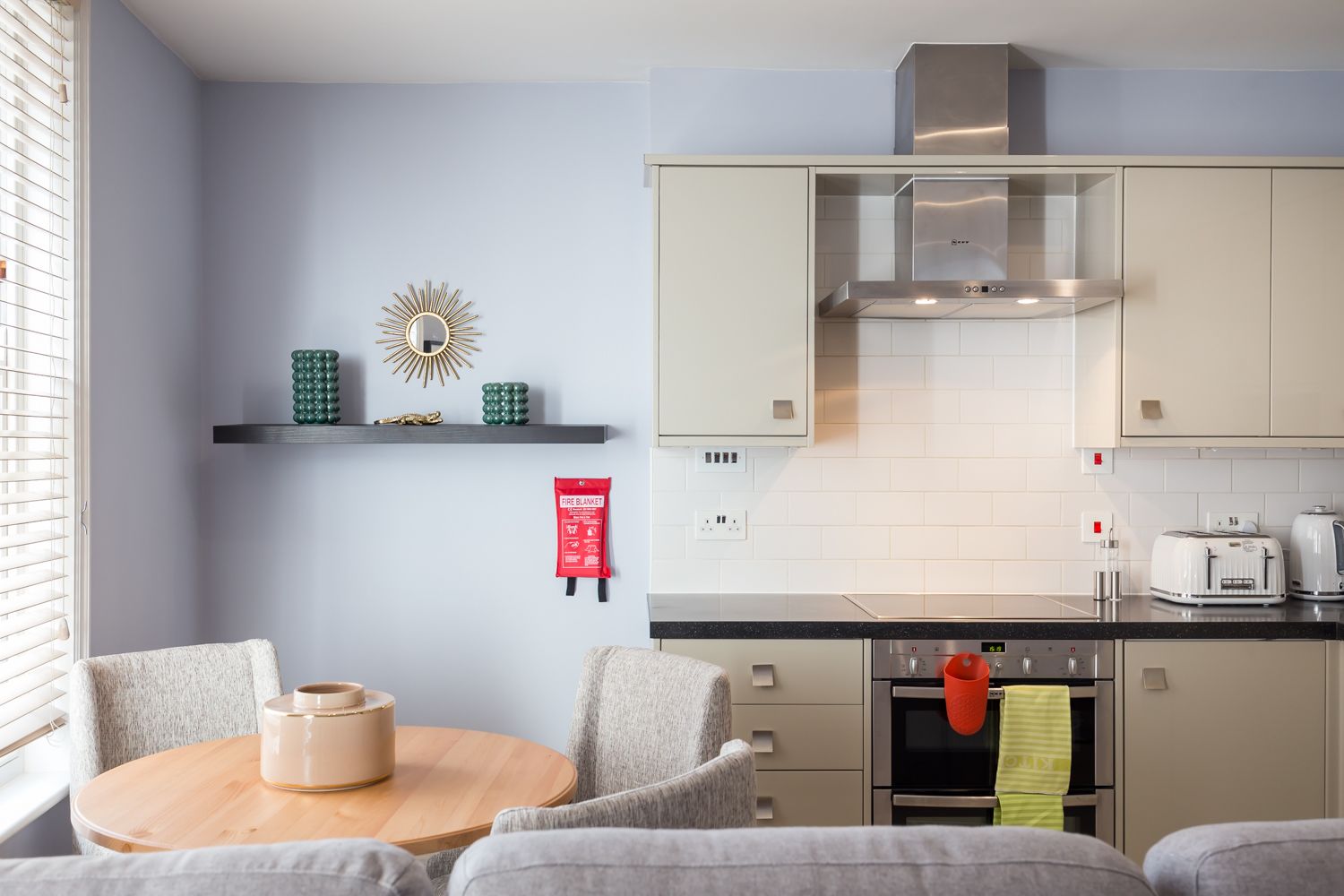A modern kitchen with a dining area, featuring a round wooden table, grey upholstered chairs, a built-in oven, and a stainless steel range hood.