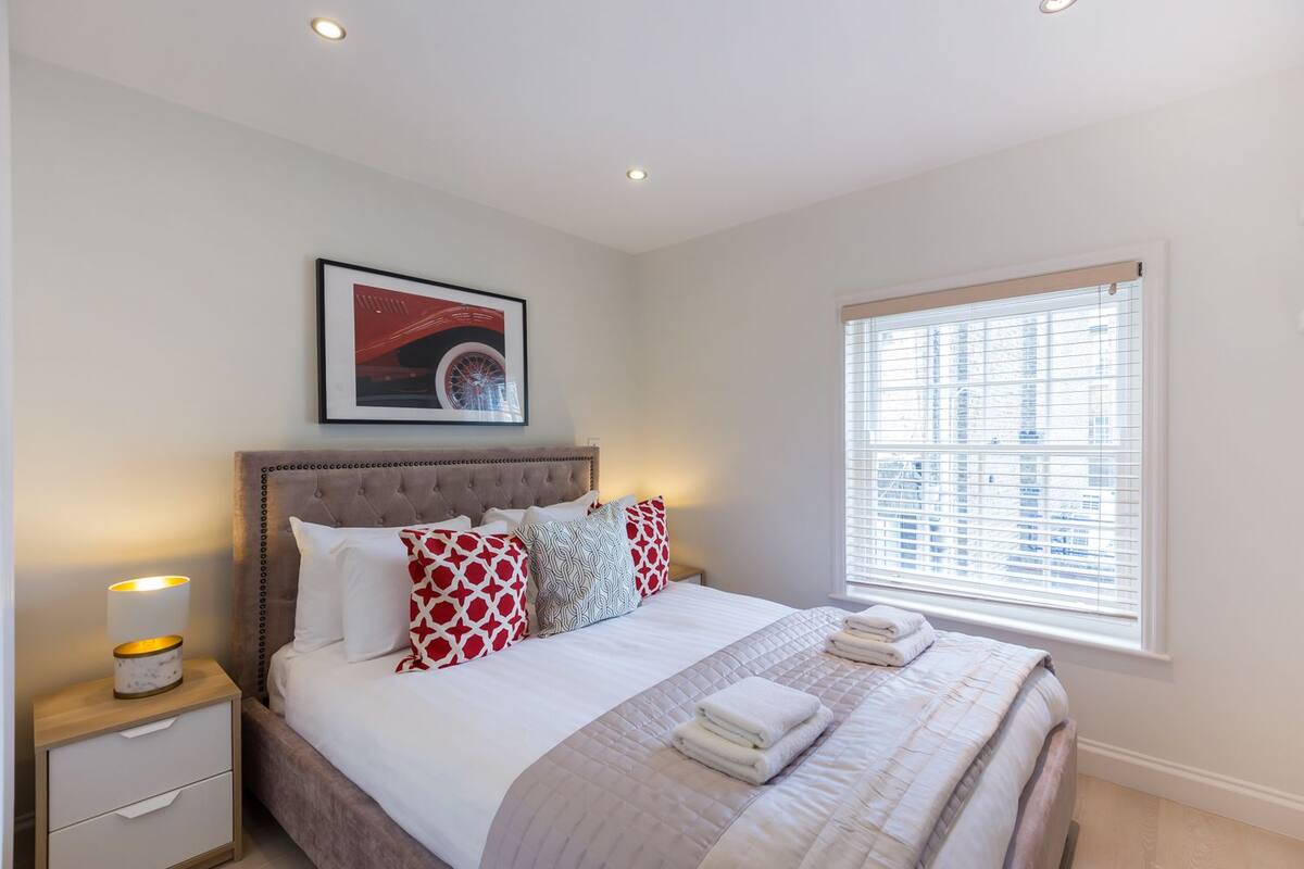 A neatly arranged bedroom with a large bed, a bedside table with a lamp, a window with blinds, and a framed picture on the wall.