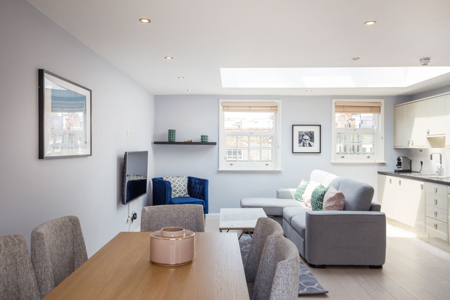 A modern living room with a wooden dining table, grey sofas, and a TV mounted on the wall.