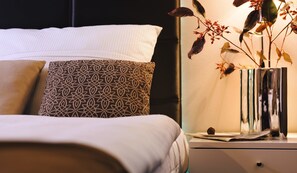 Una cama bien hecha con almohadillas estampadas, una mesita de noche con un florero de flores secas y un plato pequeño con una trufa de chocolate.
