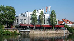 A modern building with a red awning, a reflection in the water, and a flag with the name "Eberhard". - Hotel Eberhards am Wasser (Bietigheim-Bissingen)