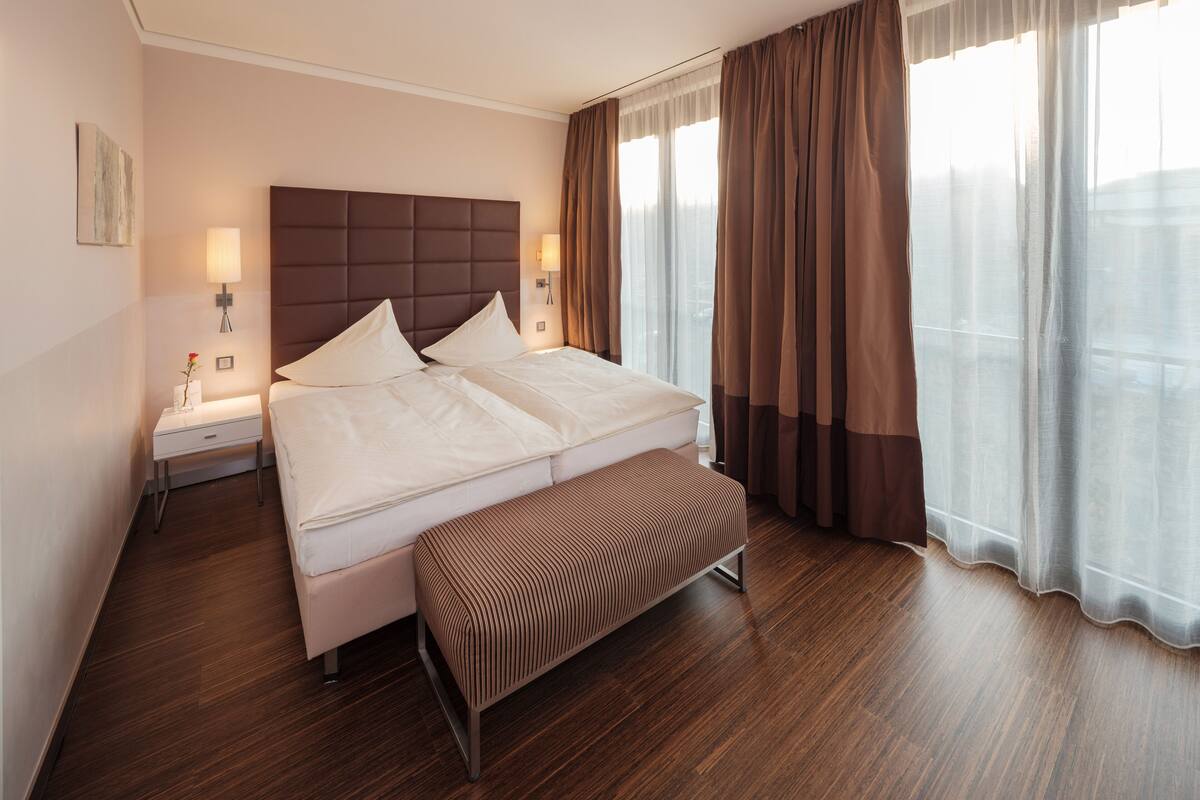 A modern hotel room with a large bed, a bedside table, a nightstand, a bench, and a view of the city through sheer curtains.