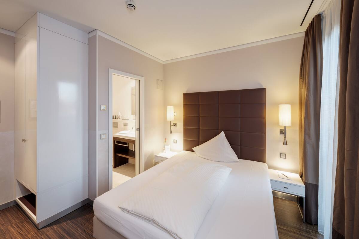 A hotel room with a large bed, bedside tables with lamps, a wardrobe, and a bathroom visible through an open door.