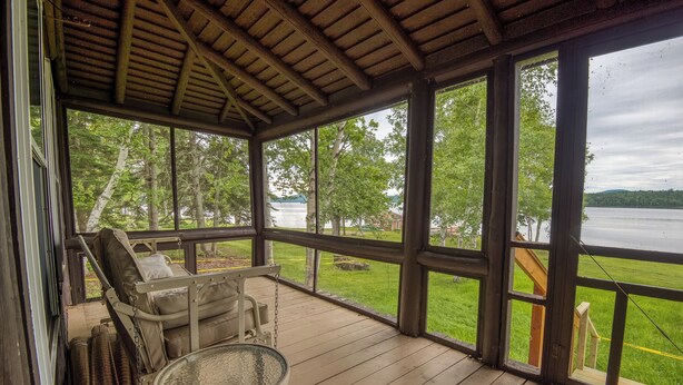 A covered porch with a view of a lake, a wooden table, and a cushioned seating area.