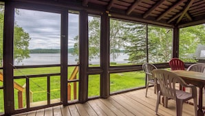 A covered deck with a view of a lake, wooden flooring, and a table with chairs.
