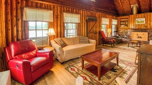 A cozy cabin interior with a leather sofa, a red armchair, a wooden coffee table, and a stone fireplace.