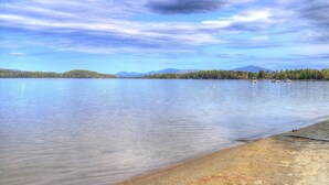 A calm lake with a clear sky, a sandy beach, and a forested island in the distance.