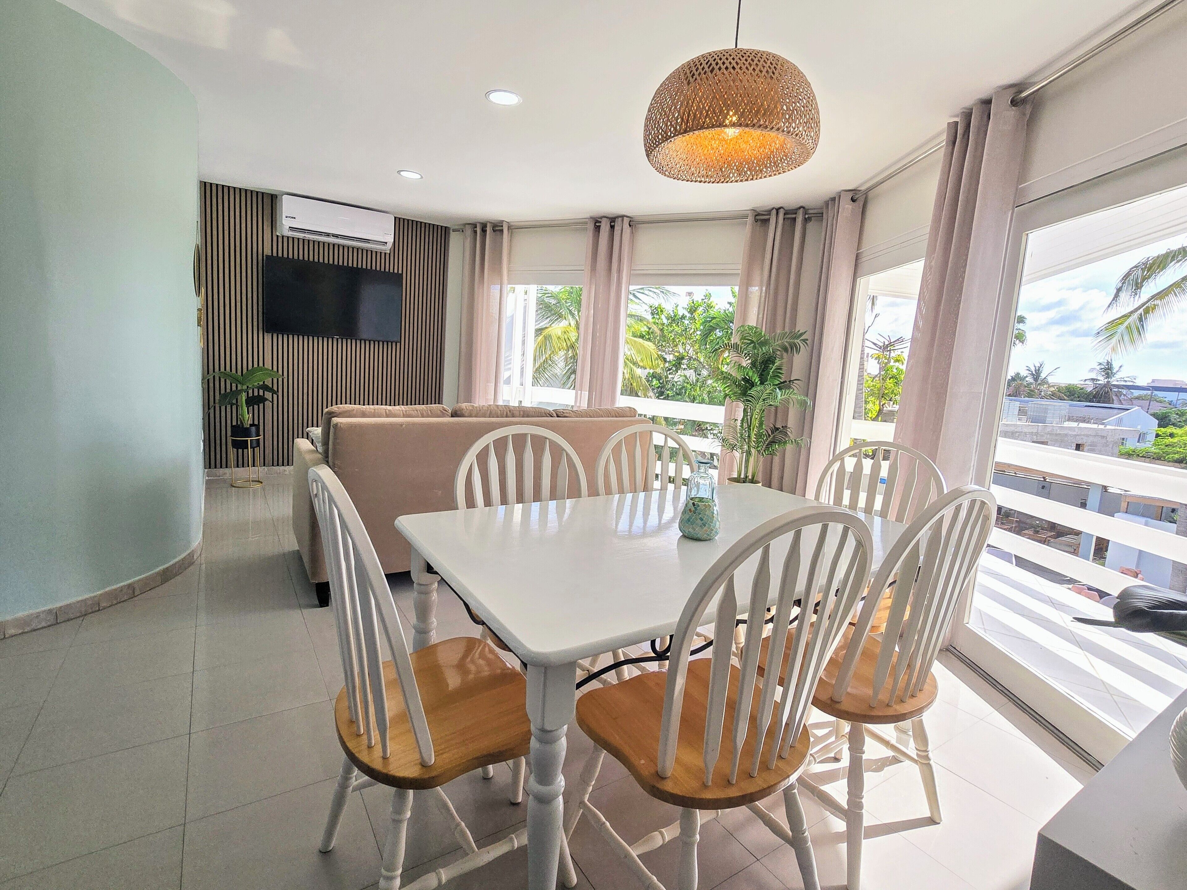 A dining area with a white table and chairs, a hanging light fixture, a sofa, and a TV mounted on the wall.