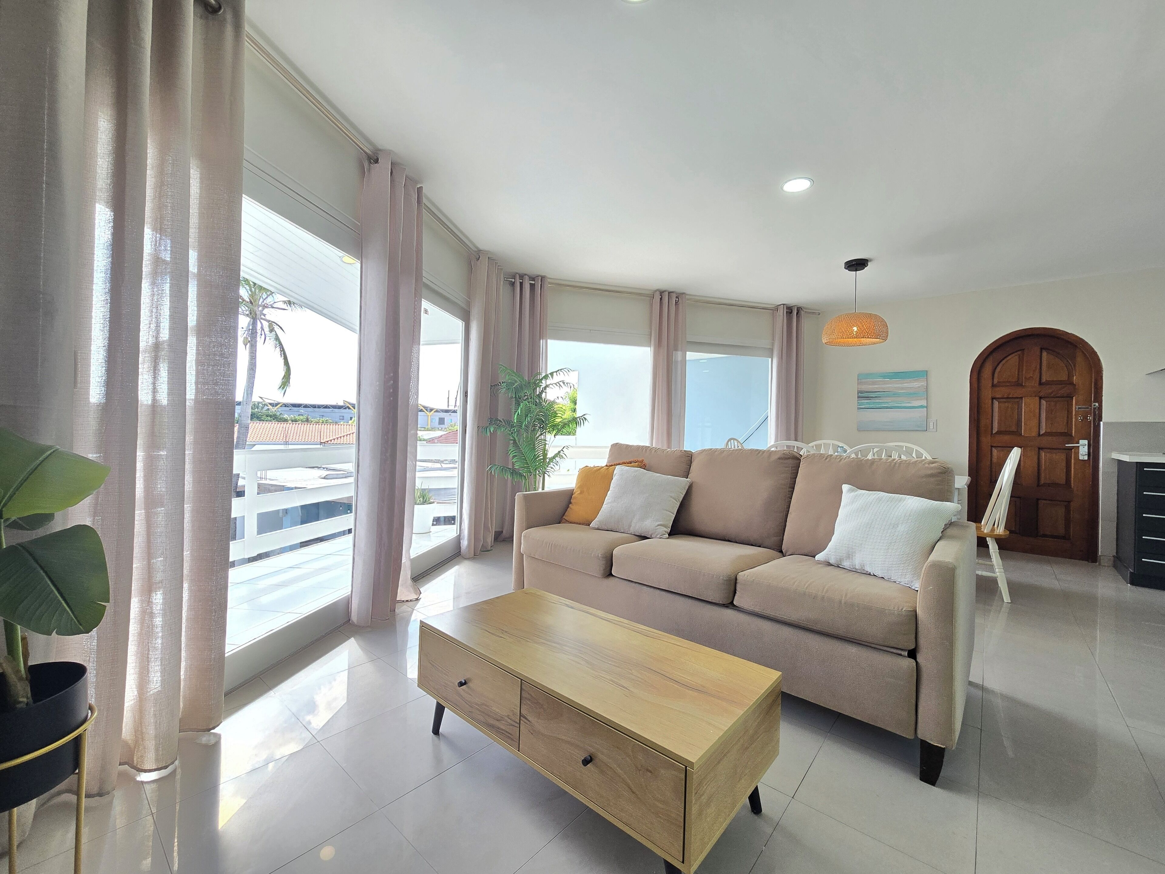 Ein modernes Wohnzimmer mit einem beigen Sofa, einem Holztisch und großen Fenstern mit Vorhängen.