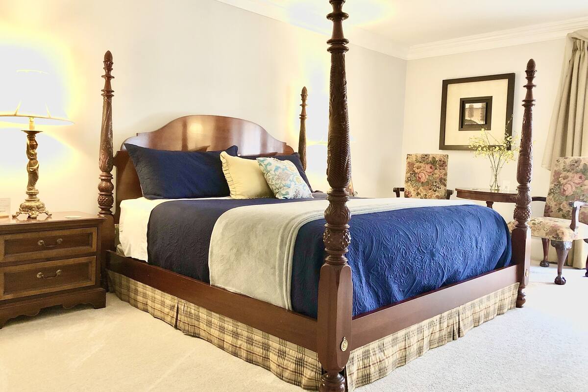 A four-poster bed with a dark wooden frame, a blue bedspread, and multiple pillows. There is a nightstand with a lamp and a small drawer. A chair and a table with a vase of flowers are visible in the background.