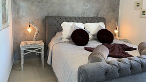 Un dormitorio con cabecera capitonada, colcha blanca, dos almohadas rojo oscuro, manta blanca y una mesita de noche con lámpara.