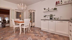 Un coin repas agrémenté d’un lustre, de chaises blanches et d’une table. On y trouve également un meuble avec des portes en verre et une étagère où sont disposées des bouteilles et des bocaux.