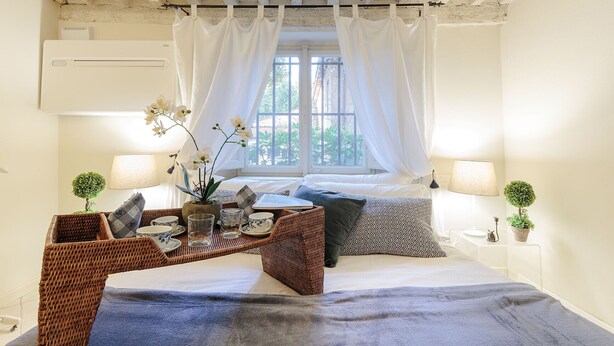 Un dormitorio con una cama, dos mesitas de noche con lámparas, una pequeña mesa con una planta y un ventanal con cortinas.