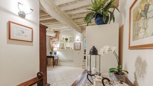 Un pasillo con un banco de madera, suelo de baldosas, una mesa con mantel a rayas, una tetera y una planta en maceta.