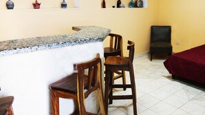 Une pièce comprenant un coin bar, des chaises en bois, un lit rouge et des étagères ornées d’objets décoratifs.