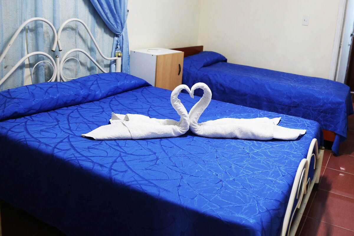 A hotel room with two beds, each covered with blue sheets. There is a heart-shaped towel arrangement on the bed in the foreground.