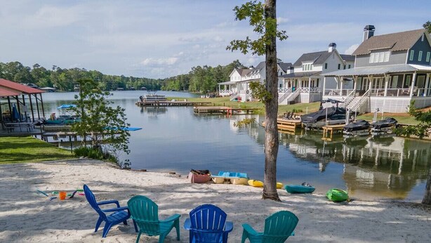 A lakeside property with a sandy beach, several Adirondack chairs, a dock with boats, and a row of houses with a lake view.