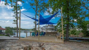 A wooden deck with outdoor seating, overlooking a lake with a house in the background.