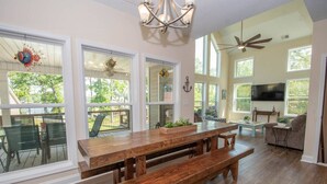 A spacious living area with a wooden dining table, benches, a ceiling fan, and large windows offering views of the outdoors.