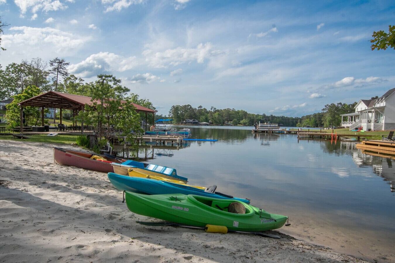 A lakeside view with a sandy beach, a covered pavilion, and a row of colorful kayaks.
