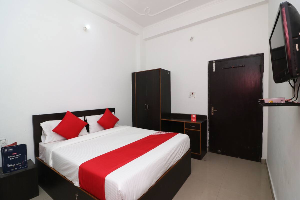 Habitación de hotel con cama, armario, televisión y una pequeña mesa con un objeto rojo encima.