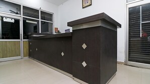 Un mostrador de recepción con acabado oscuro y detalles en forma de rombo, un ventanal con persianas y un cuadro en la pared.