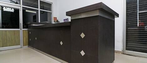 Un mostrador de recepción con acabado oscuro y detalles en forma de rombo, un ventanal con persianas y un cuadro en la pared.