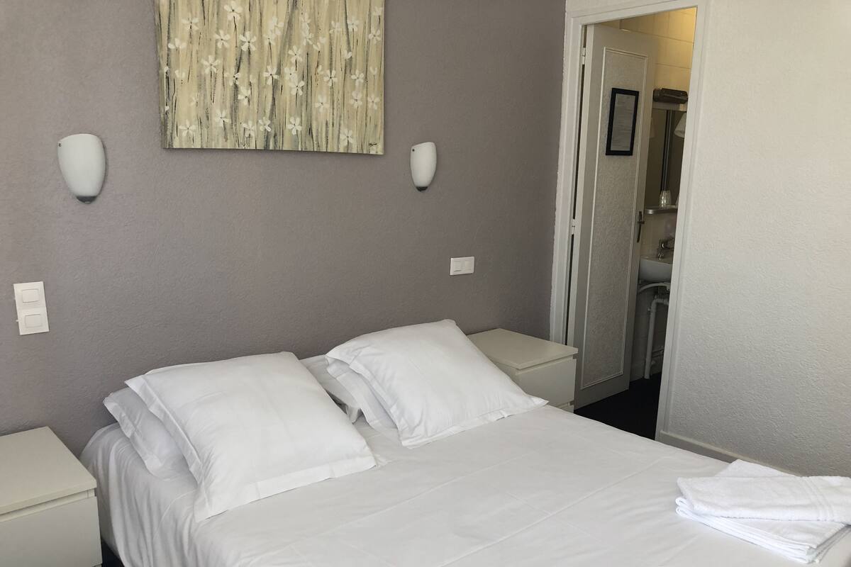 Un lit bien fait, avec du linge de lit blanc, une table de chevet et un luminaire fixé au mur.