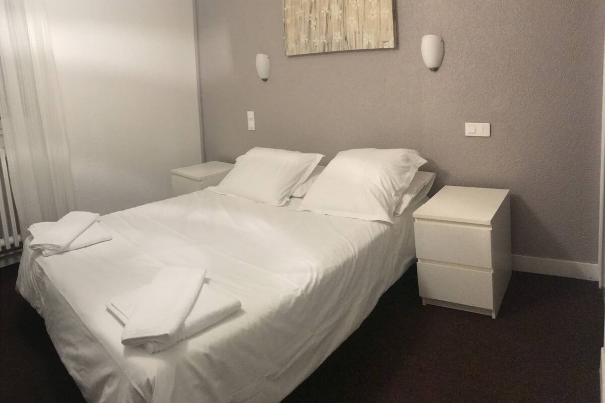 Un lit bien fait, avec du linge de lit blanc, une table de chevet, une commode et un luminaire fixé au mur.