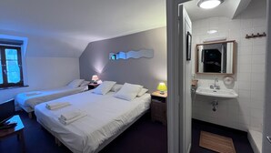 Habitación de hotel con dos camas, una mesita de noche con lámparas, un baño con lavamanos y espejo, y una ventana con persianas.