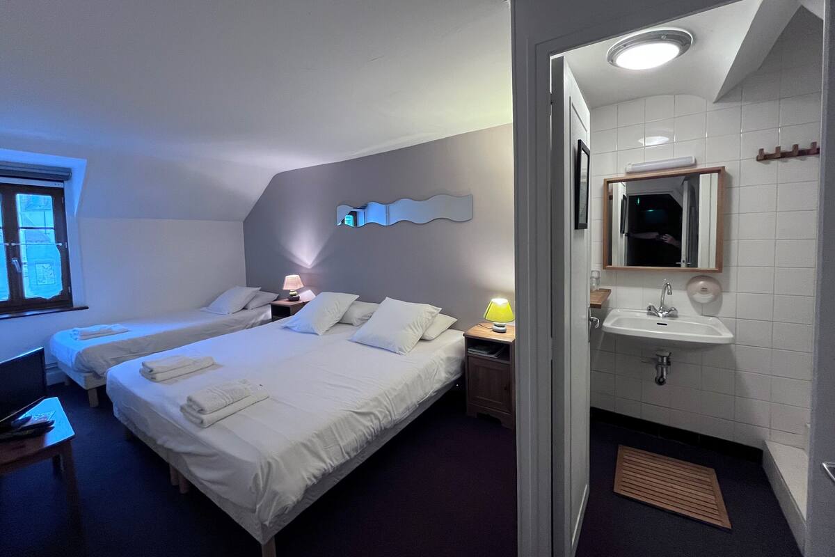 Une chambre d’hôtel avec deux lits, une table de chevet avec des lampes, une salle de bain avec un lavabo et un miroir, et une fenêtre avec des stores.