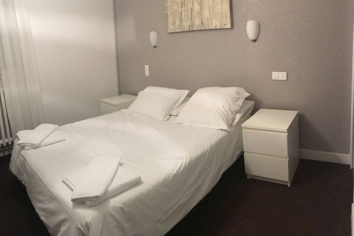 Un lit bien fait, avec du linge de lit blanc, deux tables de chevet et un luminaire fixé au mur.