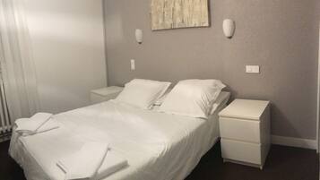 Una cama bien tendida con sábanas blancas, dos mesitas de noche y una lámpara fijada a la pared.