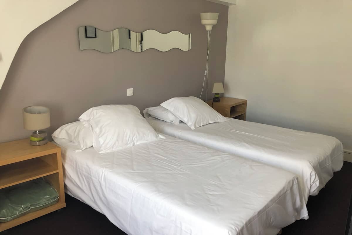 Une chambre d’hôtel avec deux lits simples, une table de chevet en bois avec une lampe, un miroir et une étagère sur laquelle se trouve un sac vert.