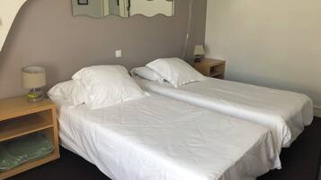 A hotel room with two single beds, a wooden nightstand with a lamp, a mirror, and a shelf with a green bag.