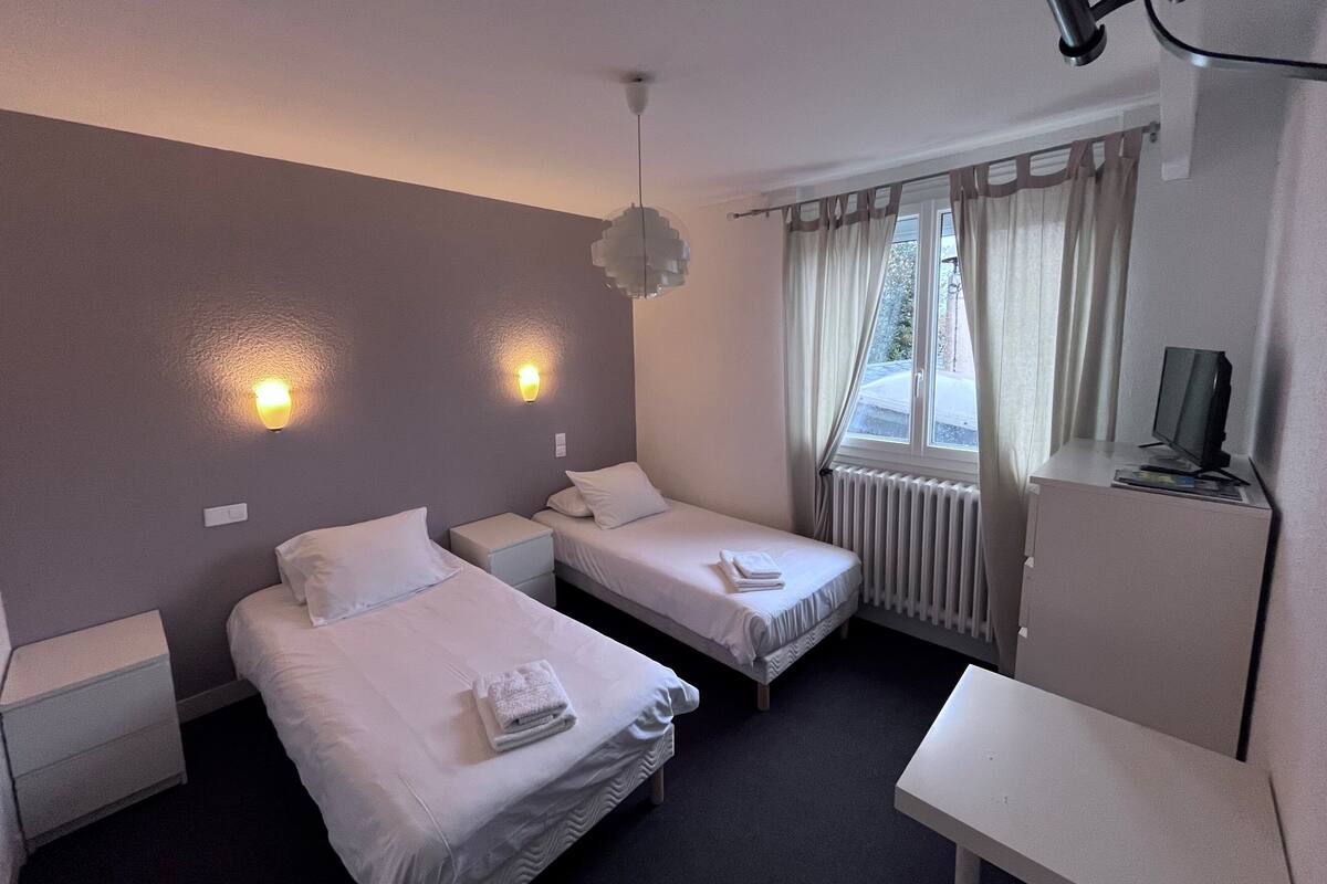 Une chambre d’hôtel avec deux lits simples, un bureau, une chaise, une télévision et une fenêtre avec des rideaux.