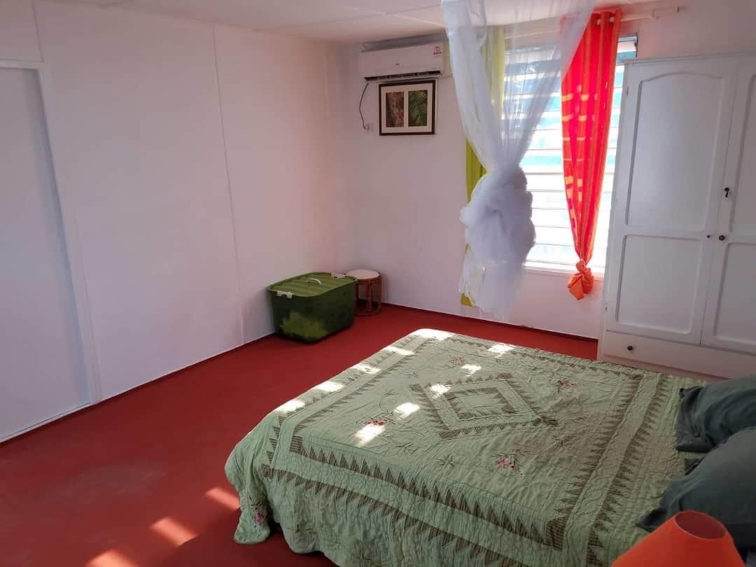 A room with a bed, a green suitcase, a white wardrobe, and a window with red curtains.
