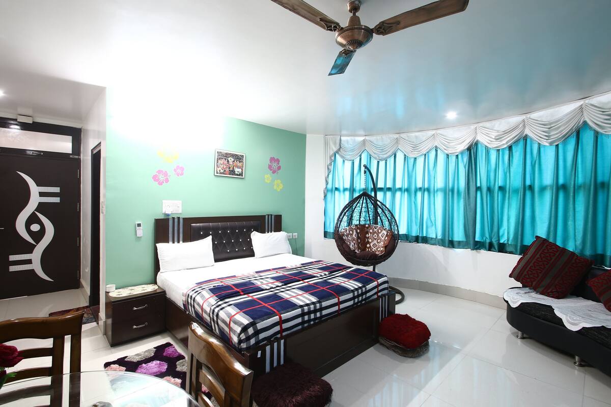 A bedroom with a bed, bedside table, hanging chair, and a window with curtains.
