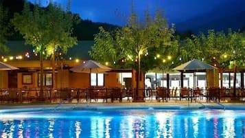 An outdoor pool area with chairs and umbrellas, surrounded by trees and a building with large windows.