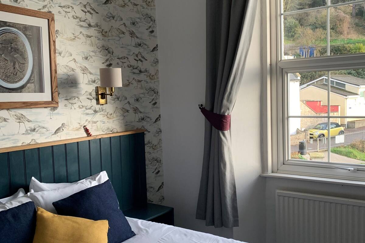 Ein Hotelzimmer mit einem Bett, einem Nachttisch, einer Wandleuchte, einem Fenster mit Vorhängen und Blick ins Freie.