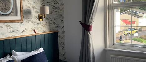A hotel room with a bed, a nightstand, a wall-mounted lamp, a window with curtains, and a view of the outside.