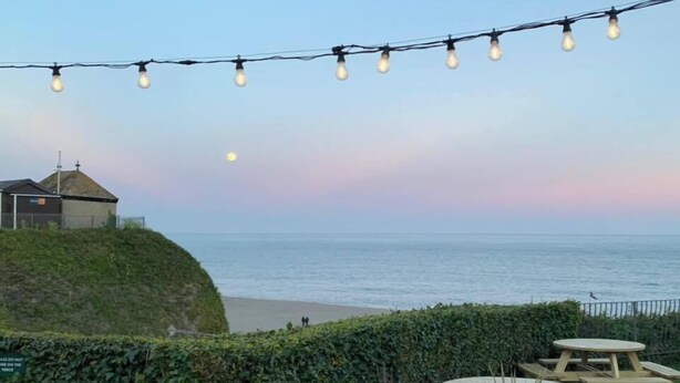 A seaside patio with string lights, wooden tables, and a view of the ocean. - Anchor Inn by Greene King Inns (Seaton)
