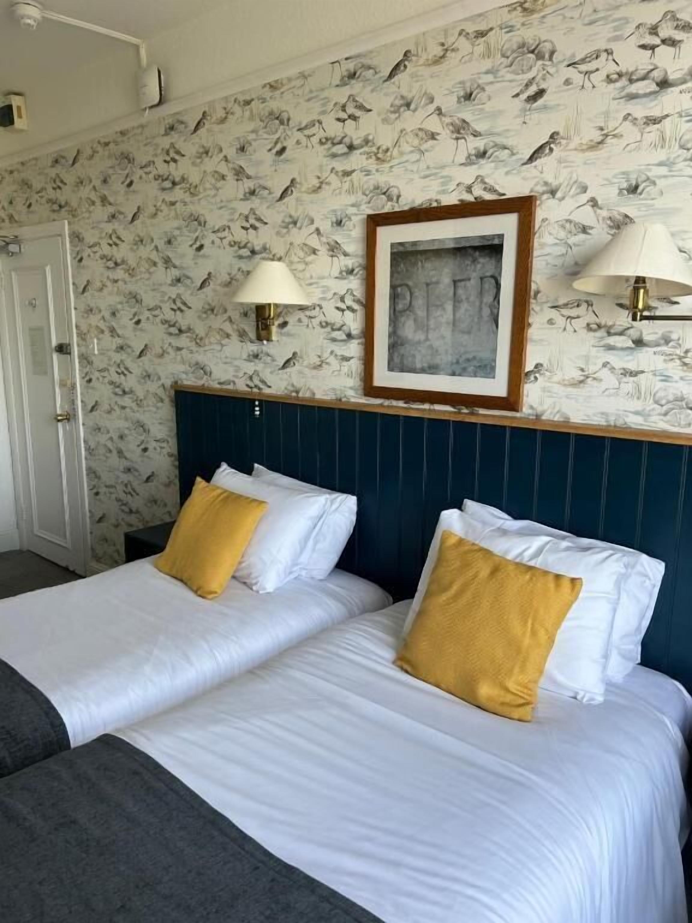 A hotel room with two beds, a framed artwork above the headboard, and wall-mounted lamps.