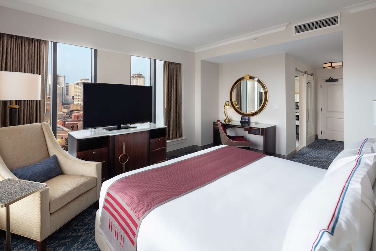 A hotel room with a large bed, a desk with a flat-screen TV, a chair, a desk lamp, a mirror, and a view of the city through large windows.