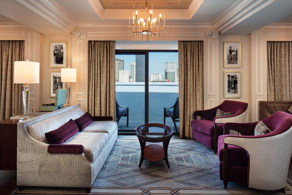 A hotel room with a sofa, armchairs, a coffee table, and a view of the cityscape through large windows.